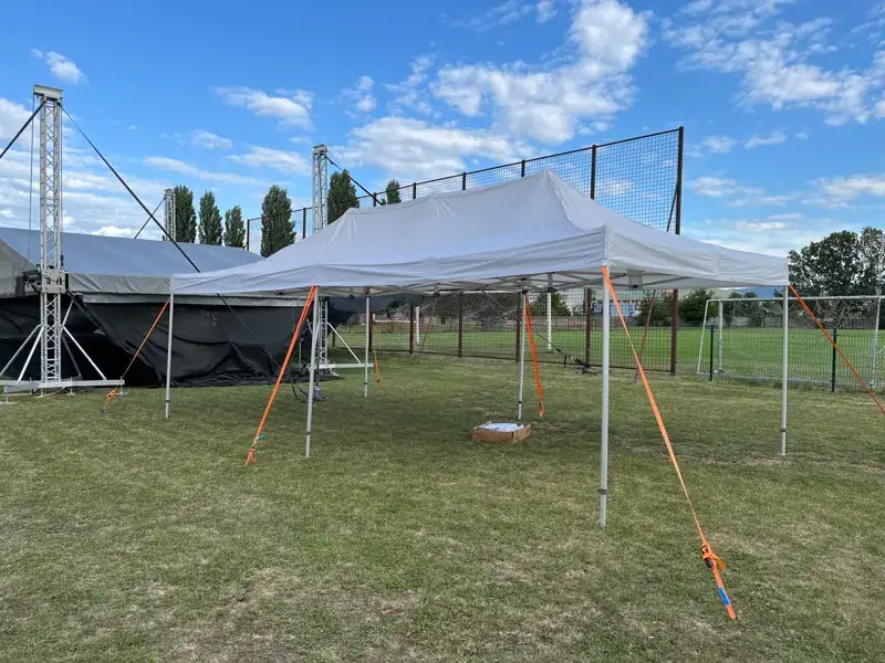 Biely nožnicový stan ukotvený popruhmi a v pozadí pódium so zastrešením zložené na zemi